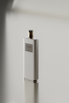 A sleek, rectangular perfume bottle with a metallic cap stands on a reflective surface. The label on the bottle reads 'DUA'. The minimalist design emphasizes simplicity and elegance.