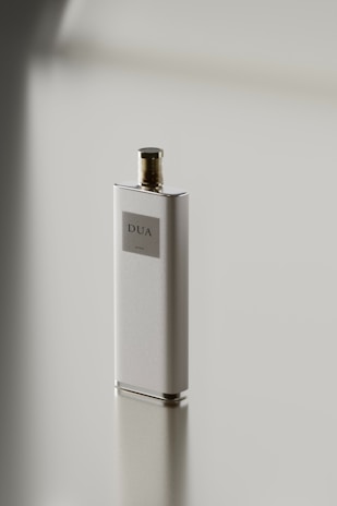 A sleek, rectangular perfume bottle with a metallic cap stands on a reflective surface. The label on the bottle reads 'DUA'. The minimalist design emphasizes simplicity and elegance.