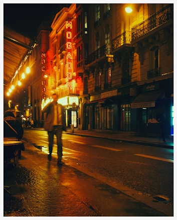 A city street at night with bright neon signs, including the word 'Mogador' prominently displayed. The street is wet, reflecting the lights from the buildings. A blurred figure walks in the foreground, and there are people sitting outside a café. The atmosphere is lively, yet calm.