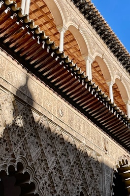 A detailed view of a historic architectural facade featuring intricate carvings and archways. The design showcases a blend of geometric and floral patterns, with a strong emphasis on symmetry and craftsmanship. The roof is adorned with a series of arches and wooden beams.