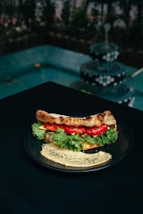 A plate with a sandwich filled with fresh lettuce, tomato slices, and possibly cheese or spread, placed on a dark table. Next to the sandwich, there is a serving of hummus garnished with black sesame seeds. In the background, there is a multi-tiered, decorative fountain with water cascading down, surrounded by greenery.