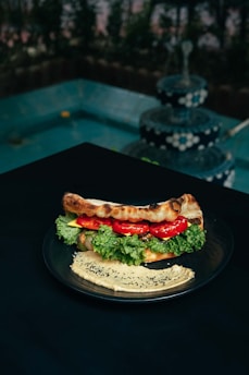 A plate with a sandwich filled with fresh lettuce, tomato slices, and possibly cheese or spread, placed on a dark table. Next to the sandwich, there is a serving of hummus garnished with black sesame seeds. In the background, there is a multi-tiered, decorative fountain with water cascading down, surrounded by greenery.