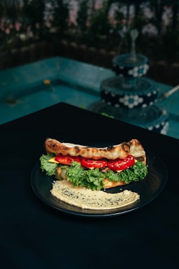 A plate with a sandwich filled with fresh lettuce, tomato slices, and possibly cheese or spread, placed on a dark table. Next to the sandwich, there is a serving of hummus garnished with black sesame seeds. In the background, there is a multi-tiered, decorative fountain with water cascading down, surrounded by greenery.