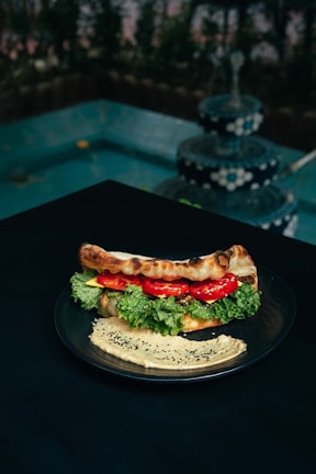 A plate with a sandwich filled with fresh lettuce, tomato slices, and possibly cheese or spread, placed on a dark table. Next to the sandwich, there is a serving of hummus garnished with black sesame seeds. In the background, there is a multi-tiered, decorative fountain with water cascading down, surrounded by greenery.