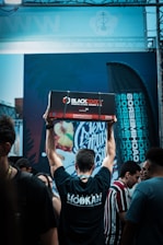 A person is standing in a crowd with their back to the camera, raising a large box labeled 'Blackcocos Premium Natural Coconut Charcoal' above their head. The person is wearing a black t-shirt with 'Hookah' written on the back. There are various people around, and a colorful graffiti-like mural is visible in the background along with a flag displaying text.