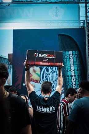 A person is standing in a crowd with their back to the camera, raising a large box labeled 'Blackcocos Premium Natural Coconut Charcoal' above their head. The person is wearing a black t-shirt with 'Hookah' written on the back. There are various people around, and a colorful graffiti-like mural is visible in the background along with a flag displaying text.