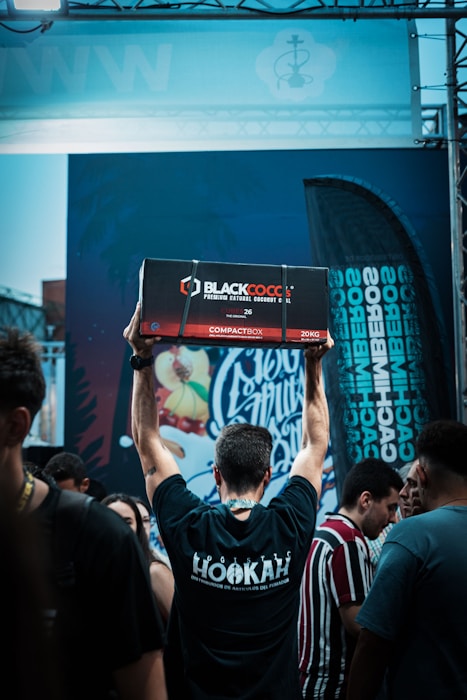 A person is standing in a crowd with their back to the camera, raising a large box labeled 'Blackcocos Premium Natural Coconut Charcoal' above their head. The person is wearing a black t-shirt with 'Hookah' written on the back. There are various people around, and a colorful graffiti-like mural is visible in the background along with a flag displaying text.
