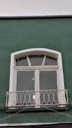 A green wall features a white-framed window with three glass panes and an arched top. Below the window, there is a small decorative metal railing with an ornate design. The paint on the window frame looks slightly aged, and some reflective light spots are visible on the glass panes.
