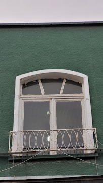 A green wall features a white-framed window with three glass panes and an arched top. Below the window, there is a small decorative metal railing with an ornate design. The paint on the window frame looks slightly aged, and some reflective light spots are visible on the glass panes.