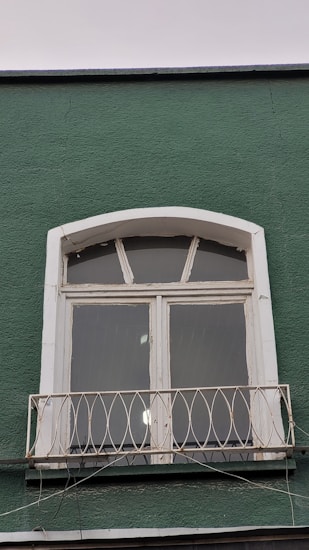 A green wall features a white-framed window with three glass panes and an arched top. Below the window, there is a small decorative metal railing with an ornate design. The paint on the window frame looks slightly aged, and some reflective light spots are visible on the glass panes.
