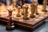 Chess pieces are arranged on a wooden, intricately carved chessboard. The focus is on the queen piece, with other pieces like pawns and knights in the blurred background. The chess pieces are made of wood in contrasting dark and light shades.