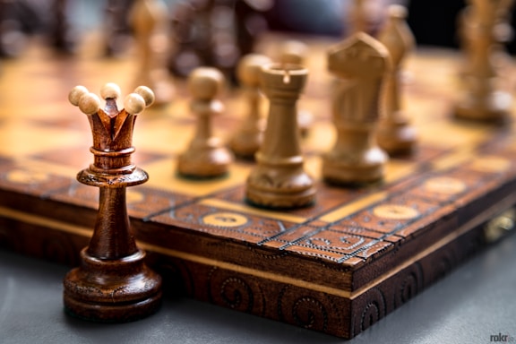 Chess pieces are arranged on a wooden, intricately carved chessboard. The focus is on the queen piece, with other pieces like pawns and knights in the blurred background. The chess pieces are made of wood in contrasting dark and light shades.