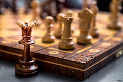Chess pieces are arranged on a wooden, intricately carved chessboard. The focus is on the queen piece, with other pieces like pawns and knights in the blurred background. The chess pieces are made of wood in contrasting dark and light shades.