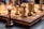 Chess pieces are arranged on a wooden, intricately carved chessboard. The focus is on the queen piece, with other pieces like pawns and knights in the blurred background. The chess pieces are made of wood in contrasting dark and light shades.