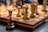 Chess pieces are arranged on a wooden, intricately carved chessboard. The focus is on the queen piece, with other pieces like pawns and knights in the blurred background. The chess pieces are made of wood in contrasting dark and light shades.