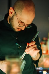 A person with glasses is focused on stirring a drink with a long metal spoon. The background is softly blurred, suggesting a bar or restaurant setting.