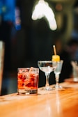 A polished wooden bar counter with three different types of glassware. In the forefront, there is a cold beverage with ice cubes in a lowball glass. Next to it, there is an empty cocktail glass and a small glass with a yellow garnish on a stick.