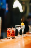 A polished wooden bar counter with three different types of glassware. In the forefront, there is a cold beverage with ice cubes in a lowball glass. Next to it, there is an empty cocktail glass and a small glass with a yellow garnish on a stick.