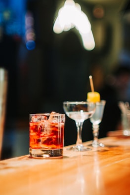 A polished wooden bar counter with three different types of glassware. In the forefront, there is a cold beverage with ice cubes in a lowball glass. Next to it, there is an empty cocktail glass and a small glass with a yellow garnish on a stick.