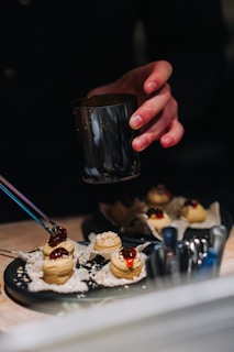 A hand holds a metallic cup over a plate of gourmet appetizers on a dark surface. The appetizers feature a textured white base topped with a spiral of mousseline and a glossy red topping. There are several metal spoons or utensils placed beside the plate, and small crumbs are scattered on the surface.