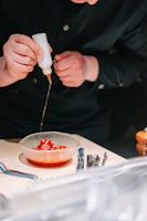 A person is carefully drizzling sauce from a squeeze bottle onto a bowl containing bright red cherry tomatoes sprinkled with sesame seeds. The table holds an assortment of utensils next to the bowl.