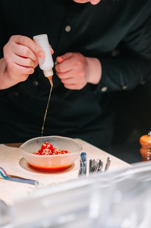 A person is carefully drizzling sauce from a squeeze bottle onto a bowl containing bright red cherry tomatoes sprinkled with sesame seeds. The table holds an assortment of utensils next to the bowl.