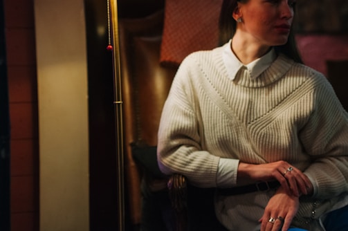 A person sits in a cozy interior wearing a white knit sweater over a collared shirt. The lighting is warm, with soft shadows. They are adorned with rings, and their hair is styled. The background includes textured materials like leather or fabric, enhancing the intimate atmosphere.