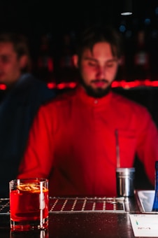 A bartender in a red shirt is standing behind a bar counter with a cocktail in the foreground, garnished with an orange slice. There is a dimly lit background with a red glow, creating a warm ambiance.
