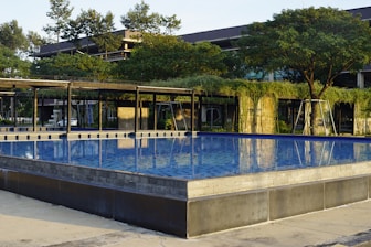 A modern swimming pool with clear blue water, surrounded by a contemporary structure adorned with greenery. Trees and overhanging plants add a natural feel to the sleek architectural design.