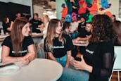 A group of young adults is engaged in a lively discussion around a table in a casual setting. They are smiling and appear to be enjoying each other's company. Many of them are wearing matching black shirts with a logo, indicating they are likely part of the same organization or event. The background has colorful graffiti or artwork, adding a vibrant touch to the ambience. Other people are visible in the background, further suggesting a social gathering or event.