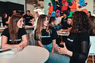 A group of young adults is engaged in a lively discussion around a table in a casual setting. They are smiling and appear to be enjoying each other's company. Many of them are wearing matching black shirts with a logo, indicating they are likely part of the same organization or event. The background has colorful graffiti or artwork, adding a vibrant touch to the ambience. Other people are visible in the background, further suggesting a social gathering or event.