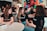 A group of young adults is engaged in a lively discussion around a table in a casual setting. They are smiling and appear to be enjoying each other's company. Many of them are wearing matching black shirts with a logo, indicating they are likely part of the same organization or event. The background has colorful graffiti or artwork, adding a vibrant touch to the ambience. Other people are visible in the background, further suggesting a social gathering or event.