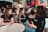A group of young adults is engaged in a lively discussion around a table in a casual setting. They are smiling and appear to be enjoying each other's company. Many of them are wearing matching black shirts with a logo, indicating they are likely part of the same organization or event. The background has colorful graffiti or artwork, adding a vibrant touch to the ambience. Other people are visible in the background, further suggesting a social gathering or event.