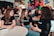 A group of young adults is engaged in a lively discussion around a table in a casual setting. They are smiling and appear to be enjoying each other's company. Many of them are wearing matching black shirts with a logo, indicating they are likely part of the same organization or event. The background has colorful graffiti or artwork, adding a vibrant touch to the ambience. Other people are visible in the background, further suggesting a social gathering or event.