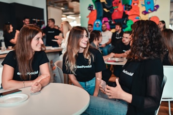 A group of young adults is engaged in a lively discussion around a table in a casual setting. They are smiling and appear to be enjoying each other's company. Many of them are wearing matching black shirts with a logo, indicating they are likely part of the same organization or event. The background has colorful graffiti or artwork, adding a vibrant touch to the ambience. Other people are visible in the background, further suggesting a social gathering or event.