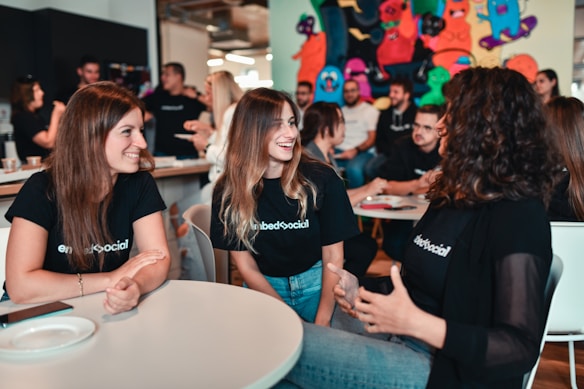 A group of young adults is engaged in a lively discussion around a table in a casual setting. They are smiling and appear to be enjoying each other's company. Many of them are wearing matching black shirts with a logo, indicating they are likely part of the same organization or event. The background has colorful graffiti or artwork, adding a vibrant touch to the ambience. Other people are visible in the background, further suggesting a social gathering or event.
