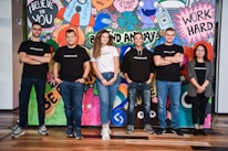 A group of six adults stand in front of a colorful mural with motivational phrases and cartoon-like drawings. They appear relaxed and confident, dressed in casual attire with matching t-shirts.