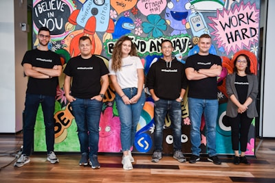 A group of six adults stand in front of a colorful mural with motivational phrases and cartoon-like drawings. They appear relaxed and confident, dressed in casual attire with matching t-shirts.