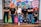 A group of six adults stand in front of a colorful mural with motivational phrases and cartoon-like drawings. They appear relaxed and confident, dressed in casual attire with matching t-shirts.