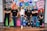 A group of six adults stand in front of a colorful mural with motivational phrases and cartoon-like drawings. They appear relaxed and confident, dressed in casual attire with matching t-shirts.