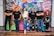 A group of six adults stand in front of a colorful mural with motivational phrases and cartoon-like drawings. They appear relaxed and confident, dressed in casual attire with matching t-shirts.