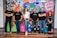 A group of six adults stand in front of a colorful mural with motivational phrases and cartoon-like drawings. They appear relaxed and confident, dressed in casual attire with matching t-shirts.