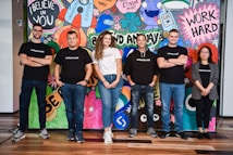 A group of six adults stand in front of a colorful mural with motivational phrases and cartoon-like drawings. They appear relaxed and confident, dressed in casual attire with matching t-shirts.