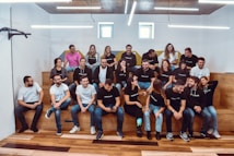 A group of people is seated on wooden steps inside a modern space with white walls, wooden flooring, and exposed ceiling lights. They are casually dressed in a mix of white and black branded t-shirts, with some individuals wearing glasses. A mix of male and female members are present, and they appear to be having a lively and cheerful discussion. The room has a minimalist design with natural light coming from a small window.
