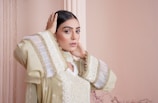 A person with long dark hair wearing an elegant, intricately designed outfit, featuring embroidered patterns and sheer fabric accents. The individual is striking a pose with one hand on their head in a softly lit setting with a neutral-colored background.