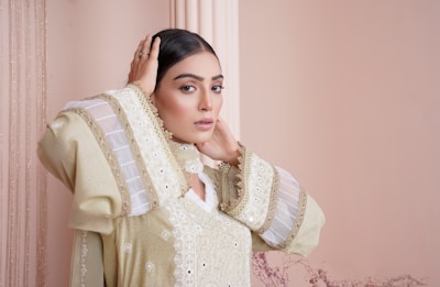 A person with long dark hair wearing an elegant, intricately designed outfit, featuring embroidered patterns and sheer fabric accents. The individual is striking a pose with one hand on their head in a softly lit setting with a neutral-colored background.