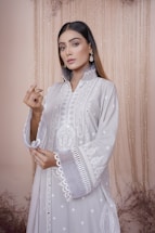 A woman dressed in an elegant, embroidered light gray outfit with intricate patterns. She wears statement earrings and has straight hair. The backdrop features a soft, beige curtain with subtle floral decor.
