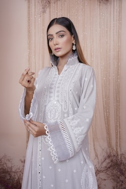 A woman dressed in an elegant, embroidered light gray outfit with intricate patterns. She wears statement earrings and has straight hair. The backdrop features a soft, beige curtain with subtle floral decor.