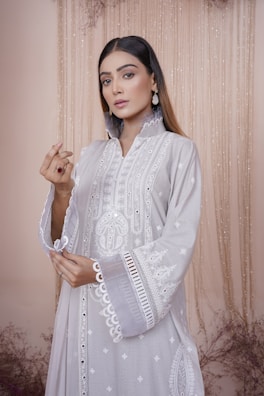 A woman dressed in an elegant, embroidered light gray outfit with intricate patterns. She wears statement earrings and has straight hair. The backdrop features a soft, beige curtain with subtle floral decor.