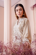 A woman wearing a beige embroidered outfit stands in front of a column with delicate dried flowers in the foreground. Her long hair is loose, and she wears gold earrings, exuding an elegant and poised demeanor.